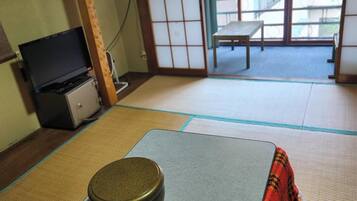 A traditional Japanese room with tatami flooring, a low table with a cushion, a television, and sliding doors leading to a balcony.