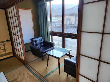 A traditional Japanese room with tatami flooring, a sliding shoji screen, a black leather sofa, a glass table, and a view of a residential area.