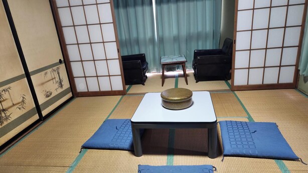 Una habitación con esteras de tatami, una mesa baja, cojines, puertas correderas y un taburete pequeño.