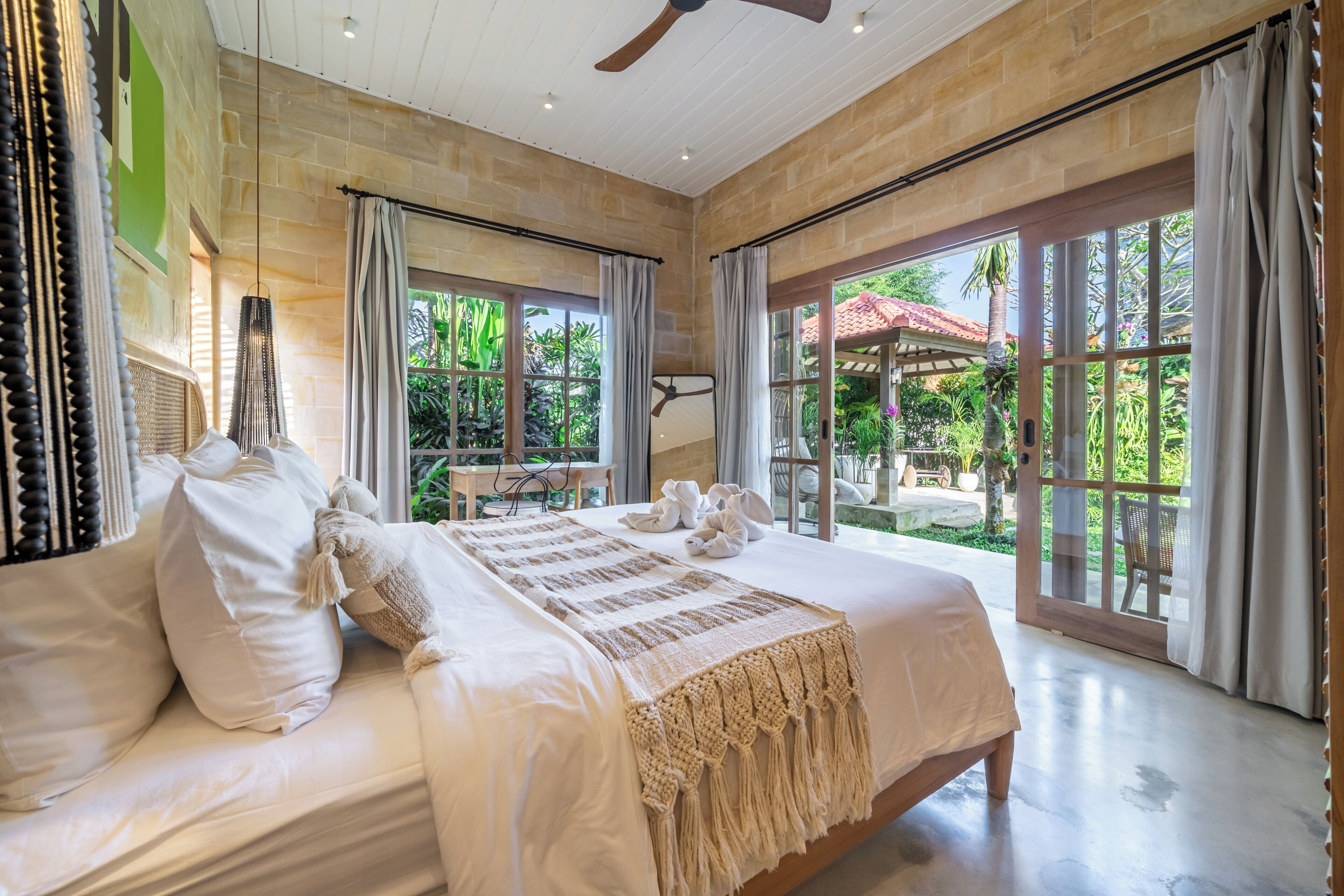 Ein geräumiges Schlafzimmer mit einem großen Bett, einem Deckenventilator und Blick auf den Garten durch große Fenster.