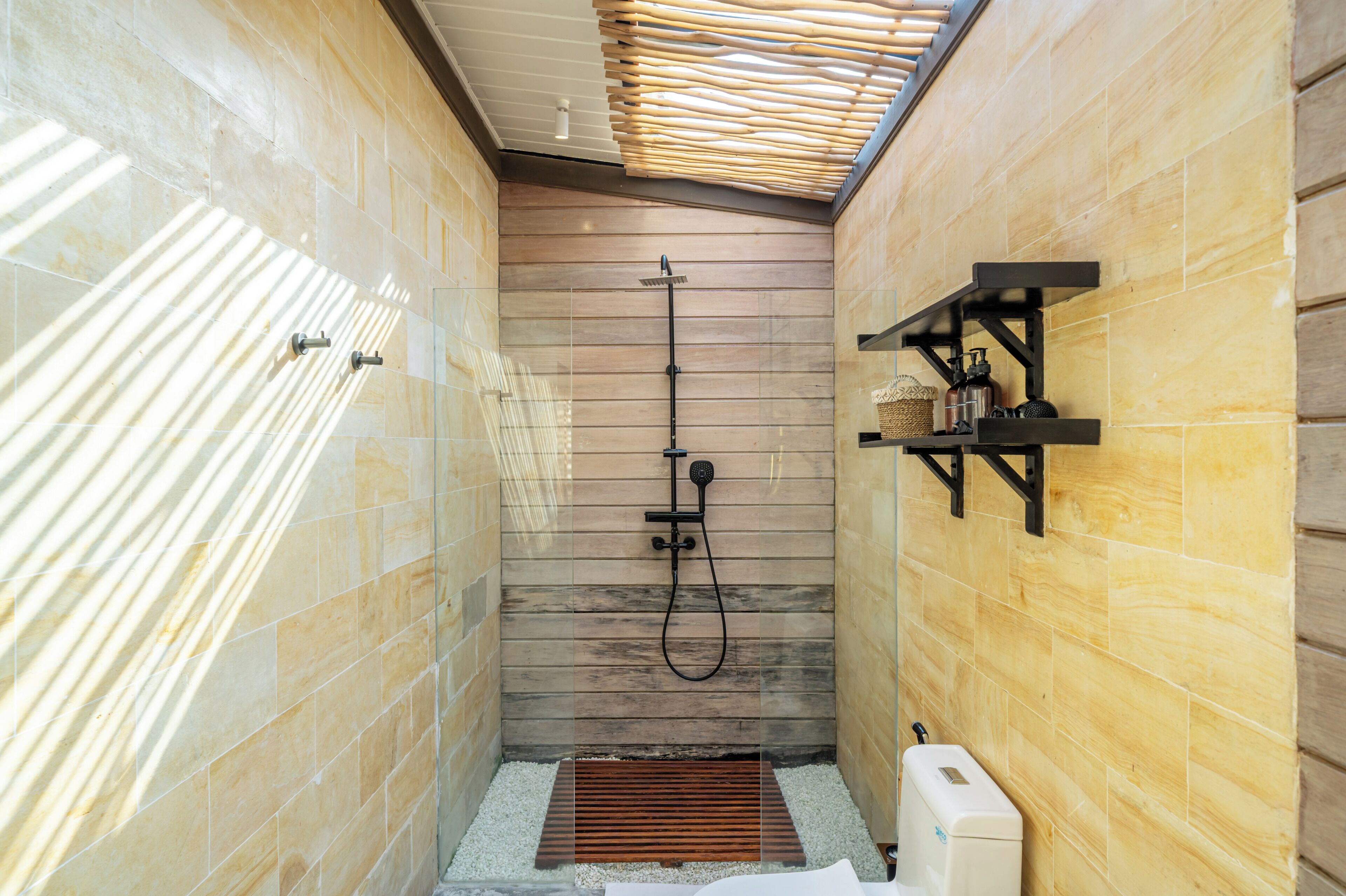 A narrow shower area with a wooden slatted ceiling, a handheld showerhead, a wall-mounted shelf with toiletries, and a white toilet.