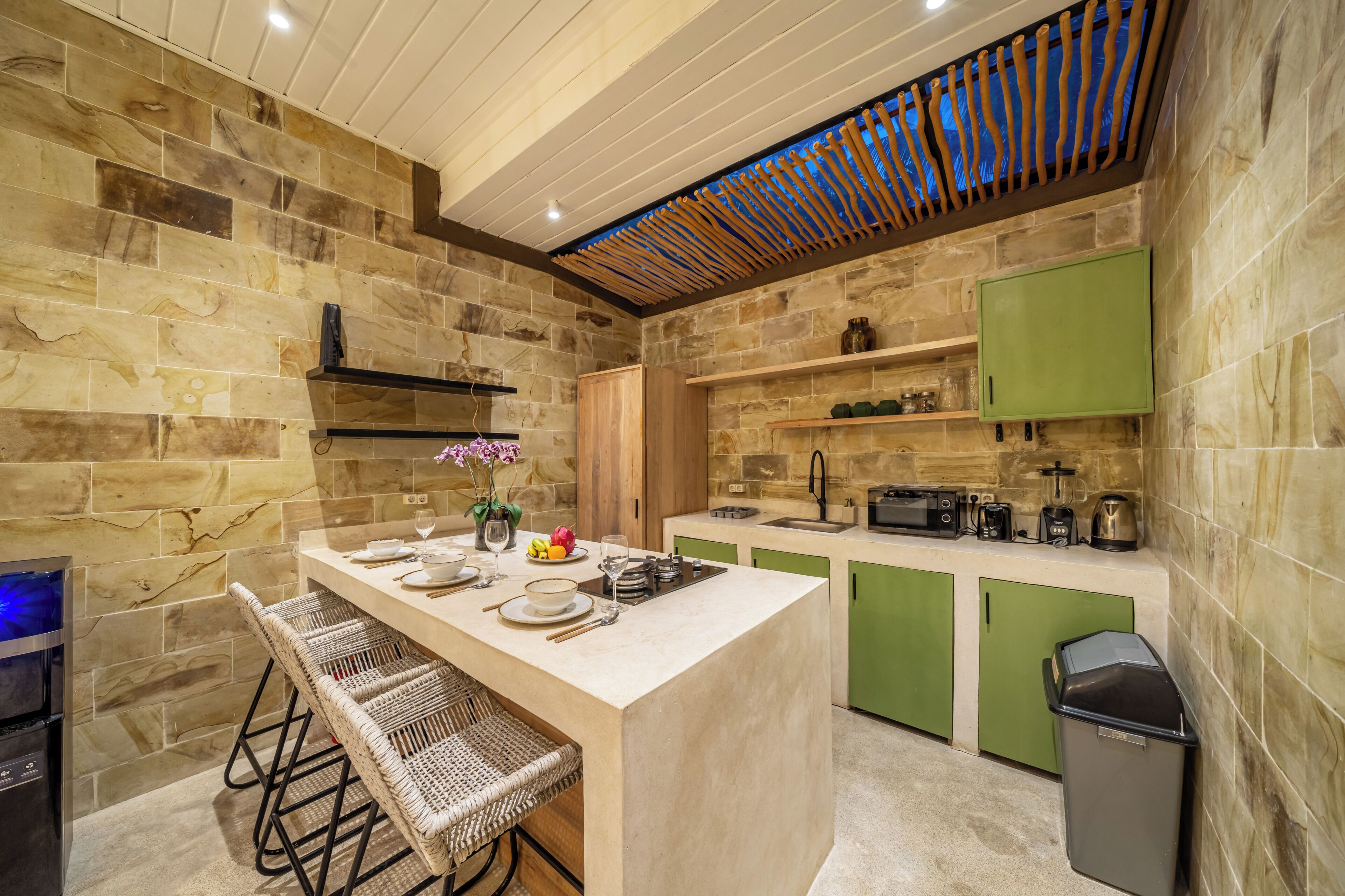Cocina moderna con salpicadero de piedra, armarios verdes y una zona de bar con sillas de mimbre.