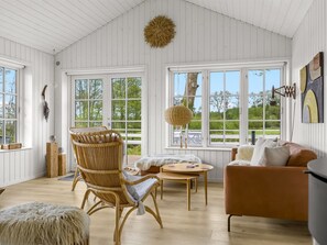 Ein geräumiges Zimmer mit hoher Decke, großen Fenstern, einem Korbstuhl, einem runden Tisch und einem braunen Sofa.
