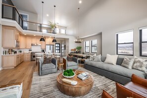 A spacious living area with a grey sofa, wooden coffee table, and a view of the outdoors through large windows. - Deer Valley East Perfect for Groups | 5BR w/ Game Room + Hot Tub (Heber City)
