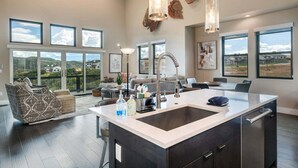 A spacious living area with a large island, modern furniture, and large windows offering views of the landscape. - Spacious Retreat | 5 King Beds | Hot Tub & Views (Deer Valley)