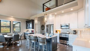 A modern kitchen with a central island, bar stools, stainless steel appliances, and a view of the outdoors. - Spacious Retreat | 5 King Beds | Hot Tub & Views (Deer Valley)
