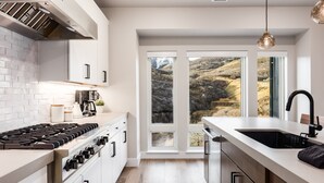 Eine moderne Küche mit Gasherd, weißen Schränken, einem großen Fenster mit Blick auf eine hügelige Landschaft und einer Pendelleuchte.