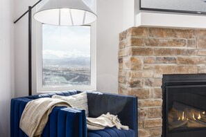 Ein Zimmer mit blauer Couch, Kamin, großem Fenster und Blick auf eine Stadtlandschaft sowie einer Lampe.