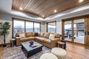A modern living room with a sectional sofa, a coffee table, and a large window offering a view of the mountains. - Luxury Ski Estate | Free Shuttle | Jordanelle Views (Heber City)