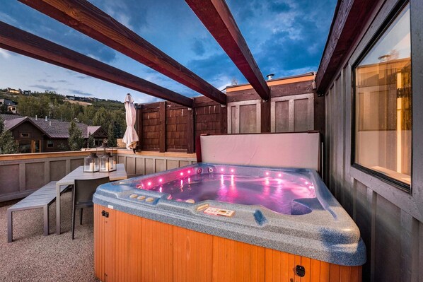 A hot tub with a wooden exterior and a stone interior, surrounded by a wooden deck and a table with chairs. - Experience luxury in this 3BR, 3.5BA loft in Deer Valley, Park City, with stunning mountain views. (Park City)