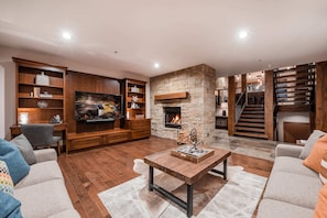 A modern living room with a stone fireplace, wooden bookshelves, a flat-screen TV, and a staircase. - Experience luxury in this 3BR, 3.5BA loft in Deer Valley, Park City, with stunning mountain views. (Park City)