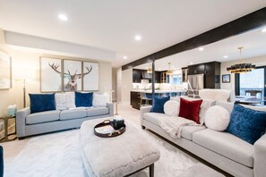Ein modernes Wohnzimmer mit einem grauen Sofa, einem weißen Sofa und einem flauschigen Ottomanen. Im Hintergrund ist ein Essbereich mit Tisch und Stühlen zu sehen.
