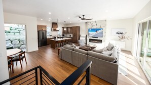 Eine moderne Küche mit Kochinsel, ein Essbereich mit Holztisch und -stühlen, ein Wohnzimmer mit Sofa und Fernseher sowie eine Treppe mit Geländer.