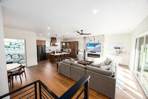 Eine moderne Küche mit Kochinsel, ein Essbereich mit Holztisch und -stühlen, ein Wohnzimmer mit Sofa und Fernseher sowie eine Treppe mit Geländer.