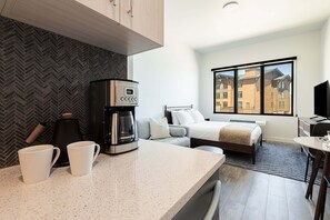 Quarto de hotel moderno com cozinha compacta, cama, janela grande e vista para edifícios.