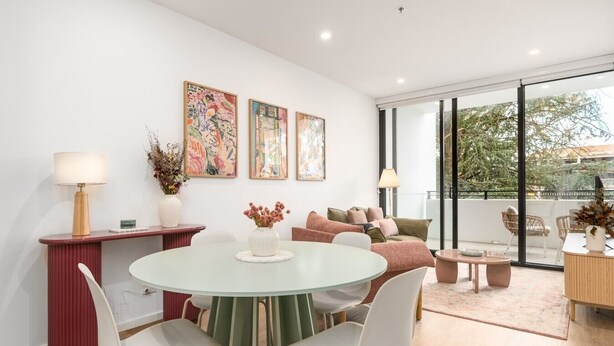 Ein modernes Wohnzimmer mit einem runden Glastisch, weißen Stühlen, einer roten Konsole und einer Couch mit Kissen. An der Wand hängen drei gerahmte Kunstwerke und ein großes Fenster mit Blick auf Bäume.