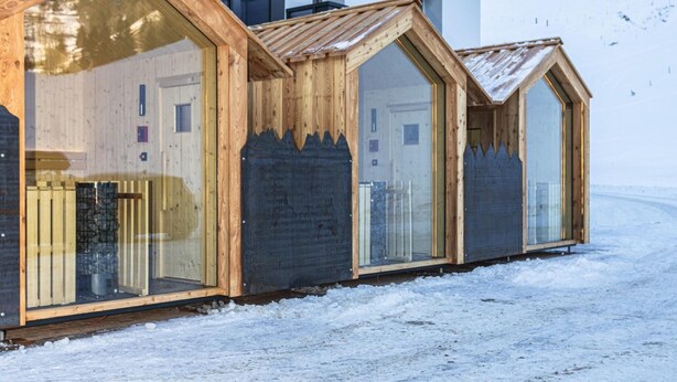 A row of wooden cabins with large windows, a white building in the background, and a snowy landscape. - Ski-in apartment with sauna in the house (Hinterglemm)