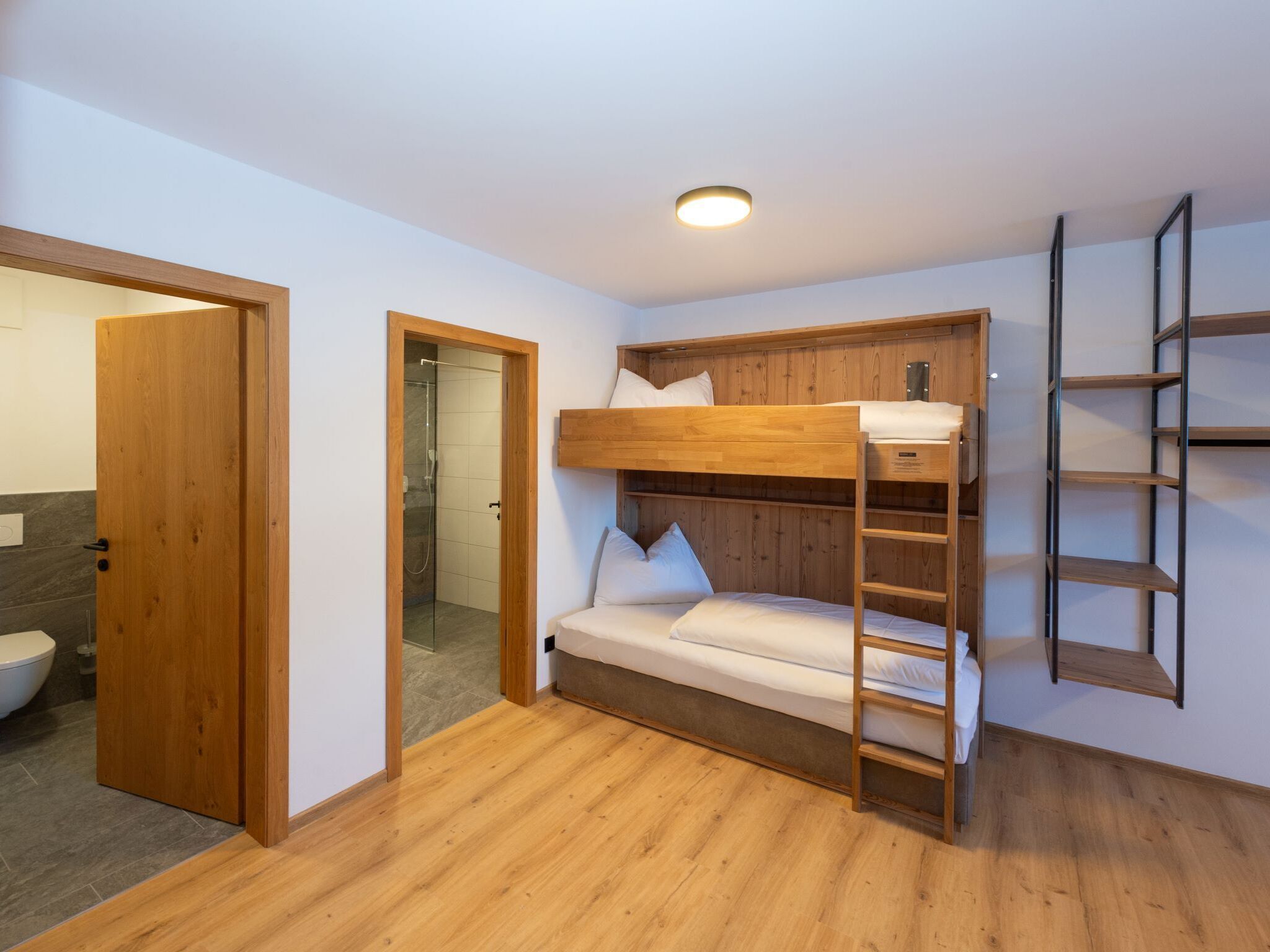 A bunk bed with a wooden ladder, a wooden shelf, and a bathroom visible through an open door.