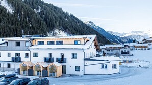Ein modernes Gebäude mit vielen Fenstern und einem geneigten Dach, umgeben von schneebedeckten Bergen und einem Parkplatz mit Autos.