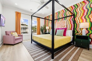 A four-poster bed with a yellow bedspread, a pink armchair, a green nightstand, and a colorful zigzag-patterned wall.
