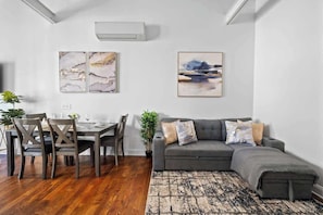 Ein modernes Wohnzimmer mit Essbereich, grauem Sofa, Holztisch und Kunstwerk an der Wand.