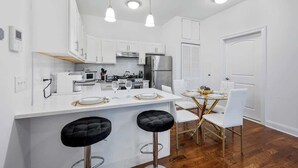 A modern kitchen with white cabinets, a stainless steel refrigerator, a dining area with a white table and chairs, and a bar stool. - 20 Min to MetLife Stadium • Perfect for FIFA Fans (Newark)