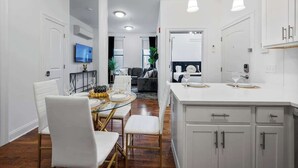 A modern kitchen with white cabinetry, a dining area with a glass table, and a living room with a sofa and TV. - 20 Min to MetLife Stadium • Perfect for FIFA Fans (Newark)
