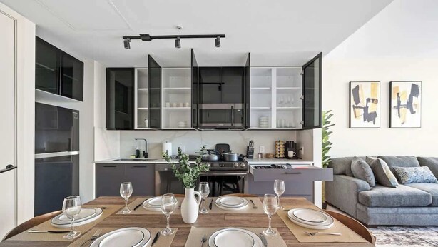 A modern kitchen with black cabinets, a dining table set for six, and a grey sofa in the living area. - Welcome to the 808 - Free parking - KING BED (East Orange)