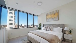 A modern bedroom with a large bed, a nightstand, a lamp, and a view of the city through large windows. - Welcome to the 808 - Free parking - KING BED (East Orange)