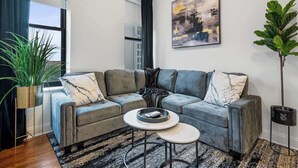 Un salon moderne avec un canapé d’angle gris, une table basse, une grande plante en pot et un tableau accroché au mur.