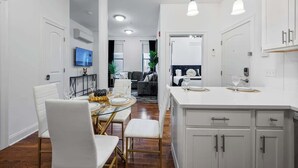 Une cuisine moderne avec des meubles blancs, un coin repas doté d’une table en verre et un salon avec un canapé et un téléviseur.