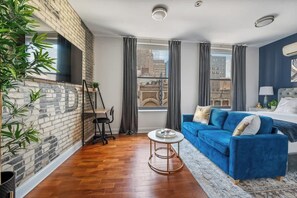 Ein modernes Wohnzimmer mit einem blauen Sofa, einem runden Couchtisch, einer Wand mit Backsteinoptik und einem großen Fenster mit Vorhängen.
