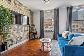 Ein modernes Wohnzimmer mit Backsteinwand, einem blauen Sofa, einem runden Couchtisch und einem großen Fenster mit Vorhängen.