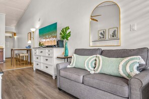 A living room with a grey sofa, a coffee table, a TV, and a large painting of a beach scene.