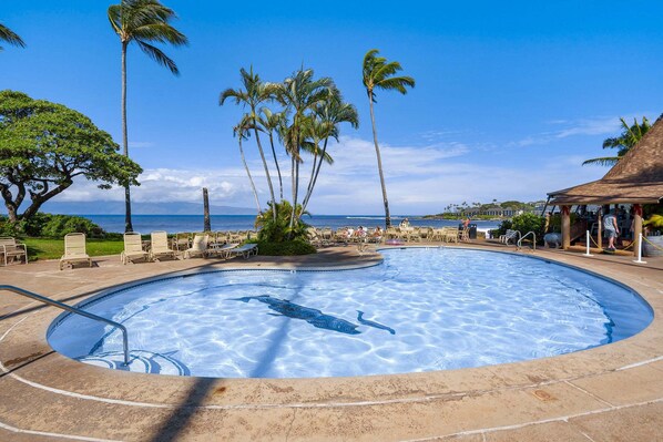 A swimming pool with a dolphin design, surrounded by palm trees and lounge chairs, with a view of the ocean and a building in the background.