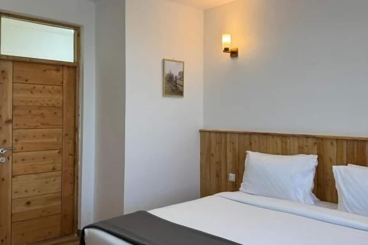 Un dormitorio con una puerta de madera, una cama con ropa de cama blanca, una pequeña lámpara de pared y un cuadro en la pared.