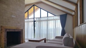 A room with a brick fireplace, a large window with curtains, and a sectional sofa. - Idan Barn Suites and Cafè (Naro Moru)