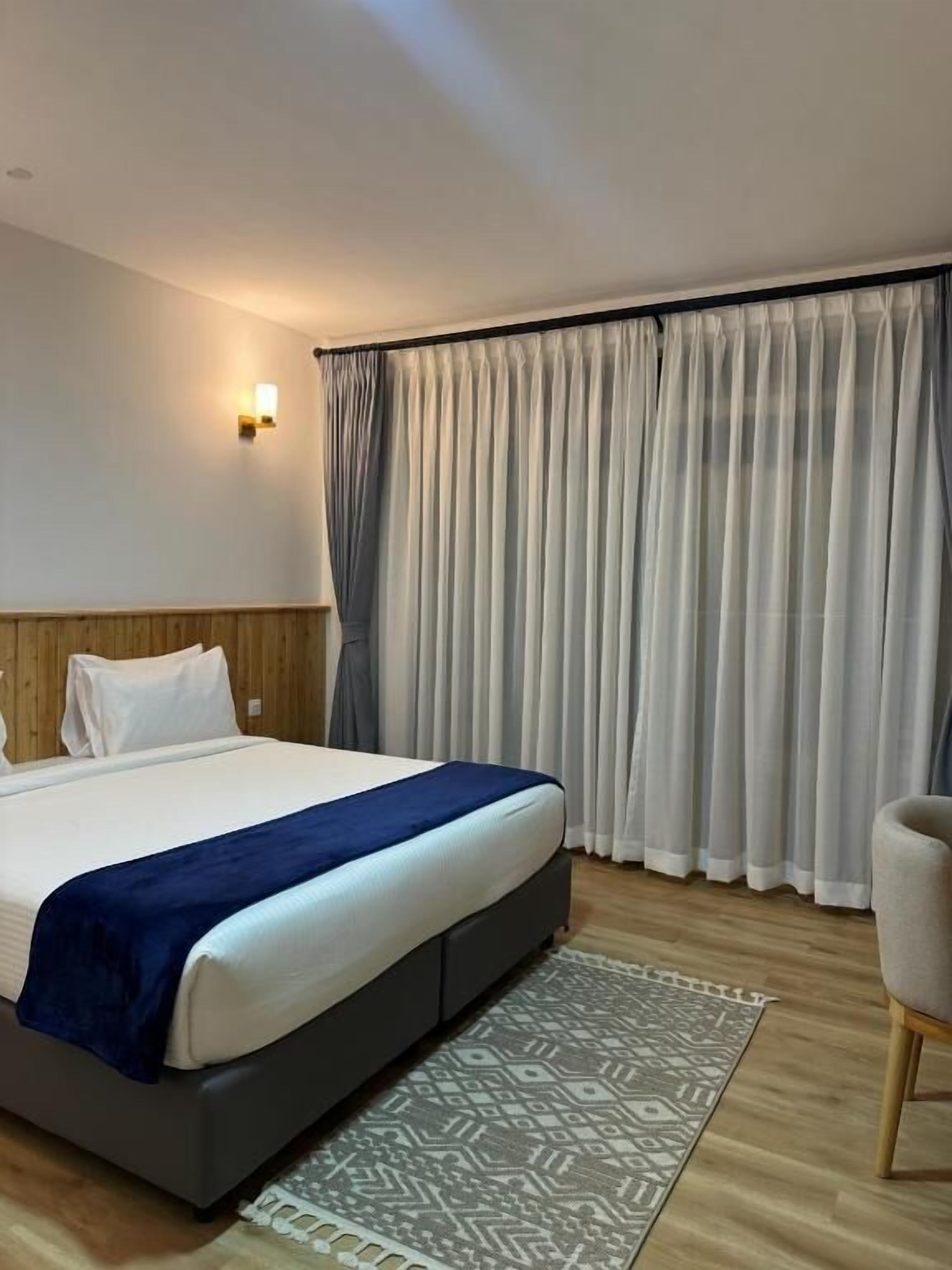 A hotel room with a bed, a nightstand, a chair, a patterned rug, and a large window with curtains.