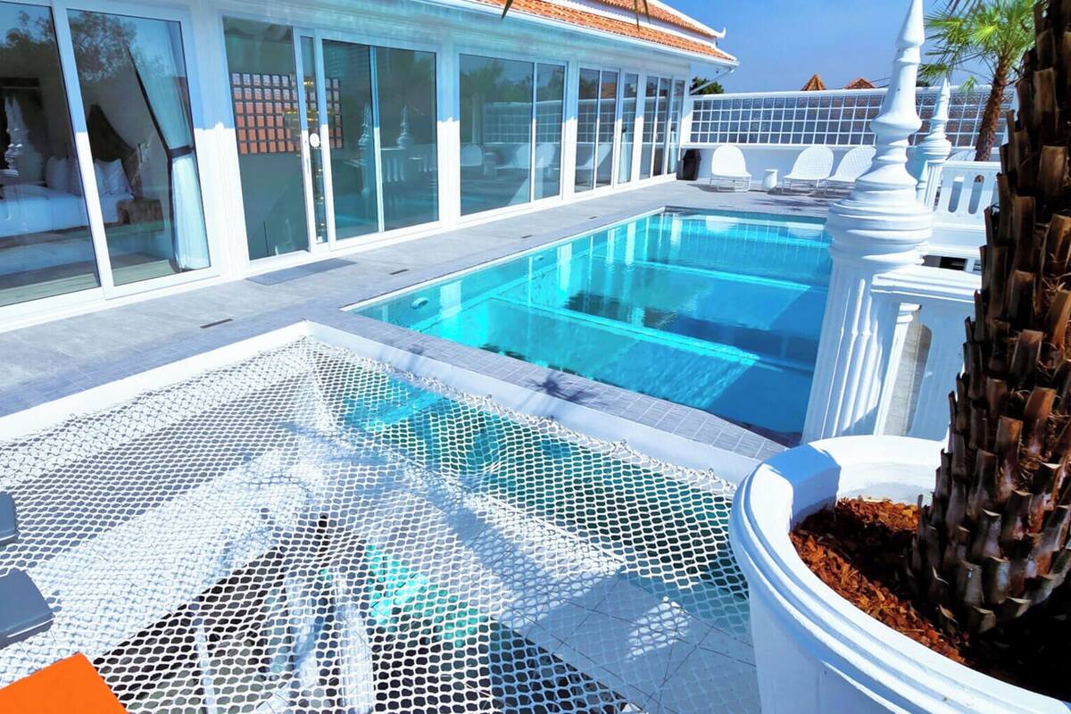 Una piscina con zona delimitada por una red, sillas reclinables blancas y una palmera.