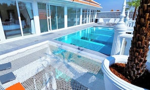 Une piscine avec une zone délimitée par un filet, des chaises longues blanches et un palmier.