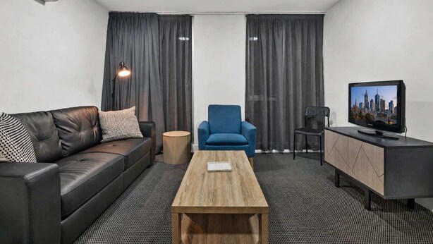 Una camera d'albergo moderna con un divano in pelle, una poltrona blu, un tavolino in legno, una TV su un mobile e una scrivania con una lampada.