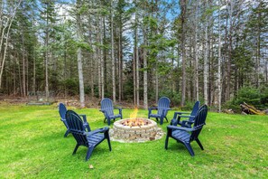 A fire pit with four Adirondack chairs arranged around it in a grassy area, surrounded by tall trees. - The Bayside Retreat-NEW! (Trenton)