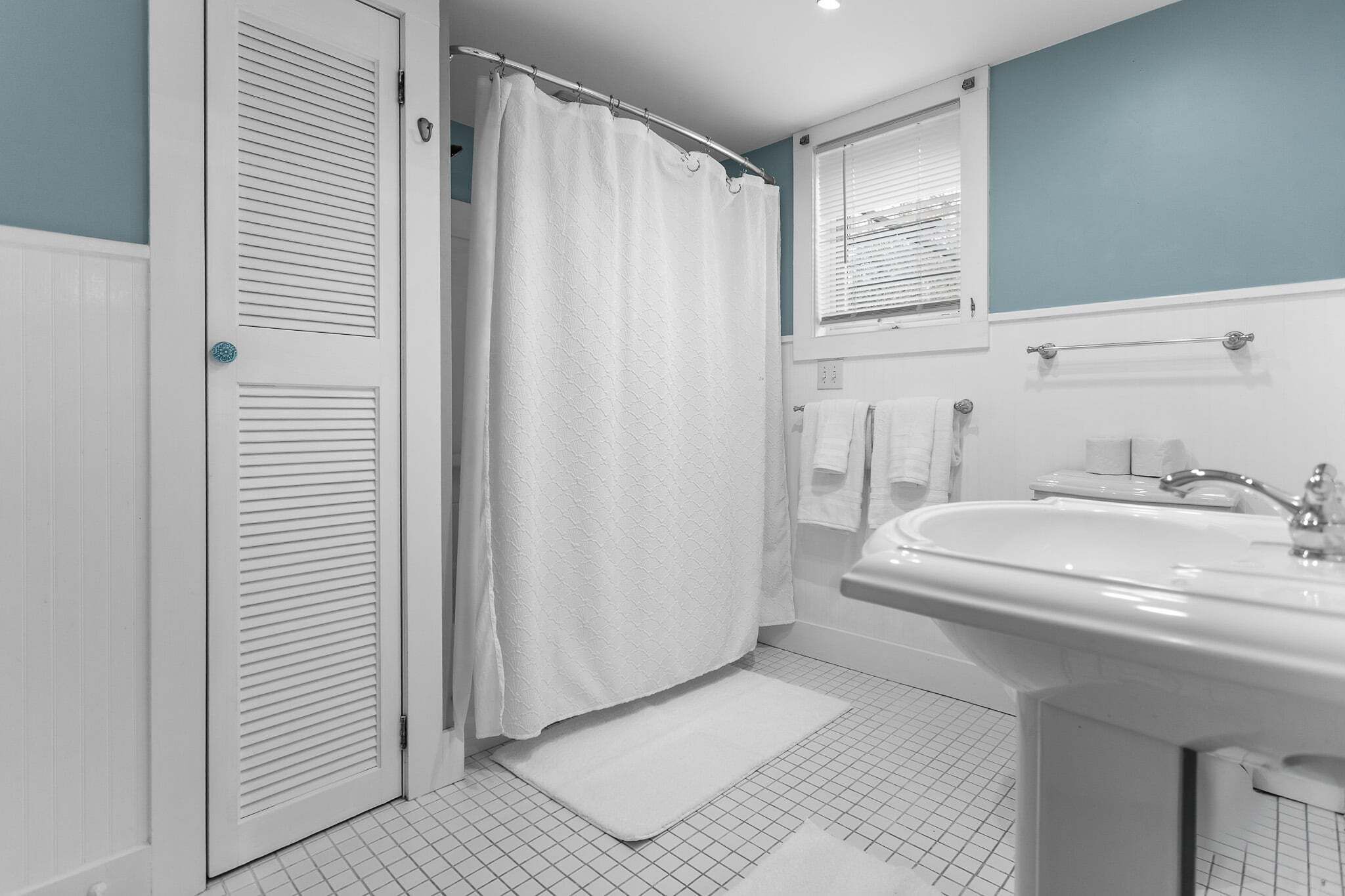 A bathroom with a white sink, a shower curtain, a white door, and a window with blinds.