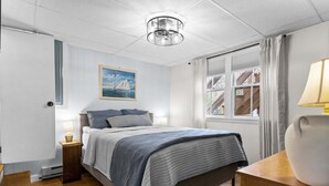 Een slaapkamer met een bed, nachtkastjes, een lamp, een schilderij, een raam met gordijnen en uitzicht op een houten terras.