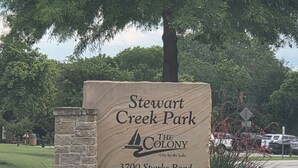 Schild für den Stewart Creek Park mit einem Steinpfeiler, gelben Blumen und einer gepflasterten Fläche.