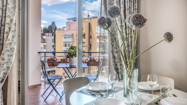 A balcony with a dining table set for two, a potted plant, and a view of buildings. - Casa Vostra Vaticano (Rome)