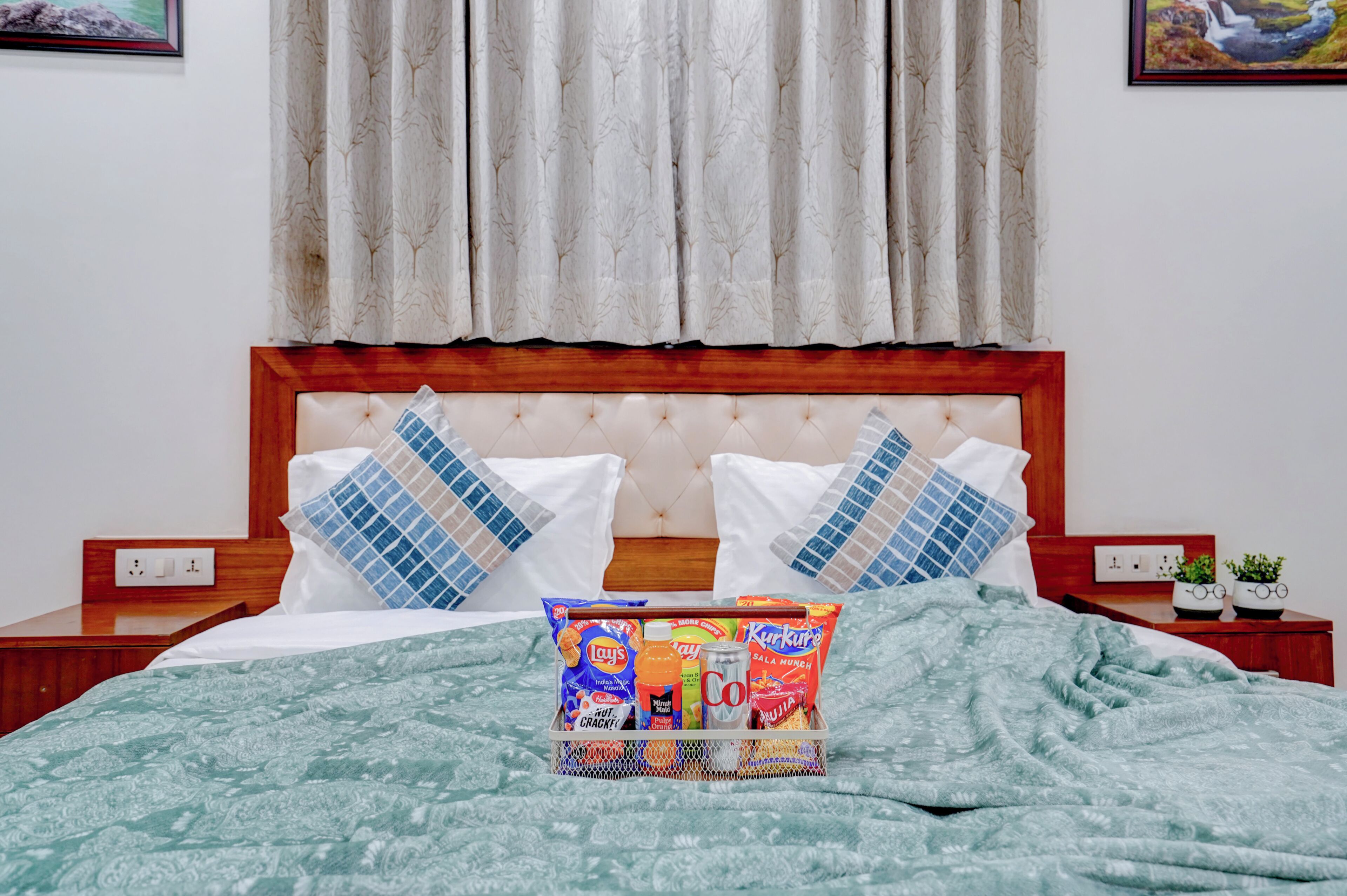 A neatly made bed with a green patterned blanket, two pillows with blue and white patterns, and a bedside table with a small plant and a framed picture.