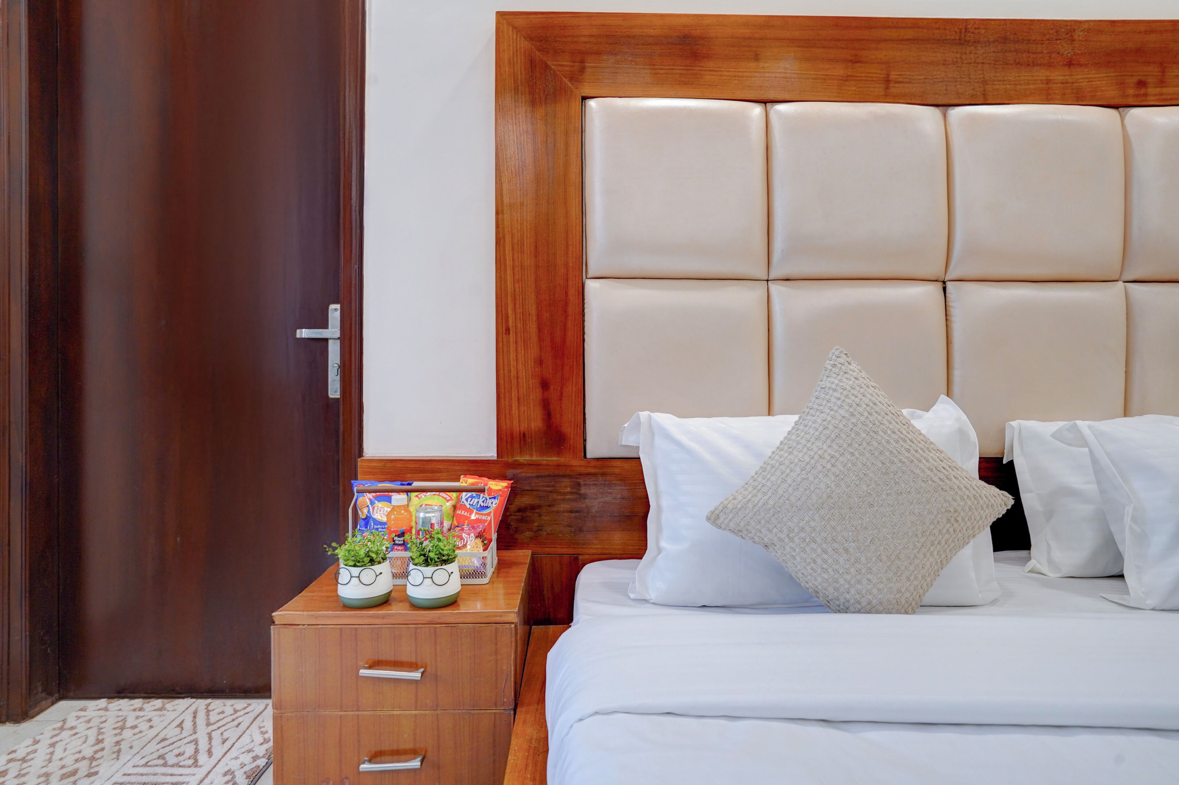 Ein Hotelzimmer mit Holz Kopfteil, einem ordentlich bezogenen Bett mit weißen Bettwäsche, einem Nachttisch mit Pflanze und Snacks sowie einer Tür mit Griff.