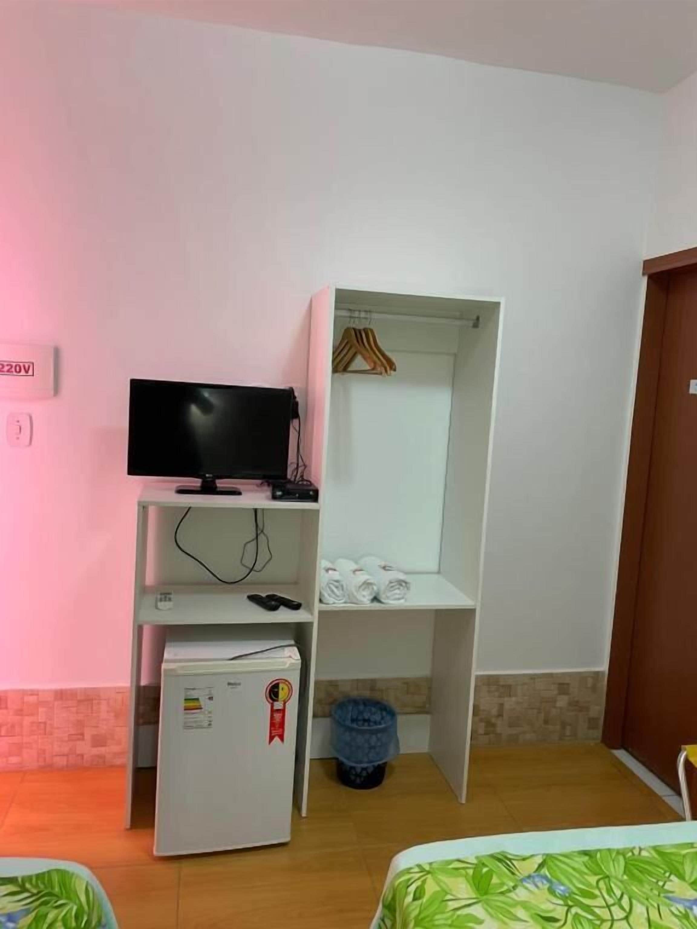 Une chambre comprenant un lit, un téléviseur posé sur un meuble, un petit réfrigérateur, une armoire avec des cintres et une poubelle.