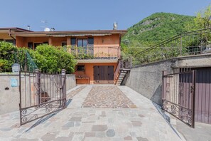 A two-story house with a stone-paved entrance, a metal gate, and a mountain backdrop. - CASA ZOLIA MENDIA, Freshness in the evening, family apartment with view. (Renzano)